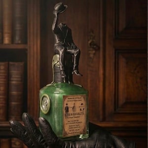 Può includere: Una bottiglia in vetro verde con etichetta Lucignolo pozione elisir di invisibilità, sormontata da una statuetta metallica scura di una persona senza testa in abito, seduta e che tiene un cappello. La bottiglia presenta un'etichetta rettangolare.