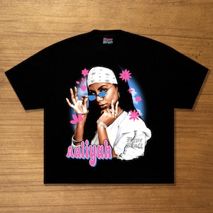 May include: A black short-sleeve t-shirt featuring a graphic print of Aaliyah wearing a white bandana and blue-tinted sunglasses. The design includes pink flowers, a blue cloud background, and the text Aaliyah in pink and blue lettering.