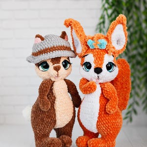 May include: Two handmade crochet plush toys. One is a brown chipmunk wearing a gray hat with large teal eyes. The other is an orange squirrel with a white belly, large teal eyes, and a small blue bow between its ears. Both have soft, fuzzy textures.