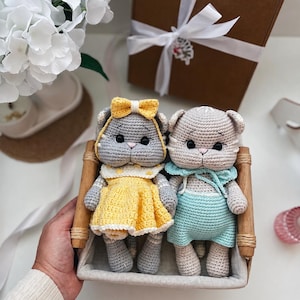 May include: Two handmade crochet cat dolls sitting in a small wooden basket. The left cat is grey with a yellow dress and matching bow. The right cat is light beige with light blue overalls and a matching collar. Both have black eyes and pink noses.