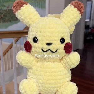 May include: A yellow plush toy with brown-tipped ears, round black eyes, a small black nose, and red circular cheeks. The toy features a stitched mouth and the text Amiigurumis printed in white across the center of the plush body.