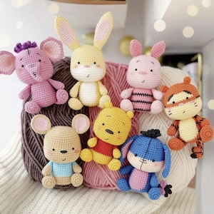 May include: A collection of six small crocheted stuffed animal figures featuring a purple elephant, a yellow rabbit, a pink piglet, a tan mouse, a yellow bear in a red shirt, and a blue donkey, all arranged against three large balls of yarn.