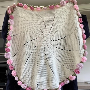 May include: A circular off-white crochet blanket featuring a radiating spiral stitch pattern. The outer edge is finished with a decorative border of small, three-dimensional crocheted roses in varying shades of light and dark pink with green leaves.