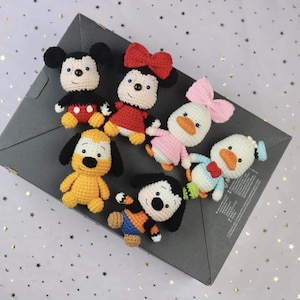 May include: A collection of five small, handmade crochet dolls featuring cartoon-inspired characters. The set includes figures with black, red, yellow, pink, and blue yarn details, each with a round head, stitched facial features, and small limbs.