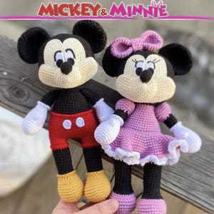 Op de afbeelding: Twee handgemaakte gehaakte poppen van Mickey en Minnie Mouse. Mickey draagt een rode broek met witte knopen en gele schoenen. Minnie draagt een paarse jurk met een bijpassende strik en paarse schoenen. De tekst MICKEY & MINNIE staat bovenaan.