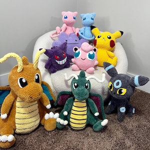 May include: A collection of ten handmade crochet plush toys featuring various colorful creatures. The group includes a large tan dragon, a green dragon, a yellow electric mouse, a purple ghost, a pink round creature, and several smaller figures.