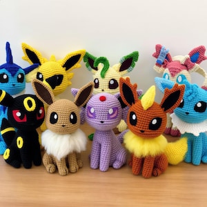May include: A collection of nine handmade crochet plush toys representing various fictional creatures. The figures feature diverse colors including black, yellow, brown, purple, and blue, with distinct ear shapes, fluffy neck ruffs, and embroidered eyes.