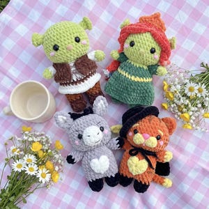 May include: Four handmade crochet plush toys including a green ogre in a brown vest, a green character with red hair in a green dress, a gray donkey with a white heart, and an orange cat with a black hat and a yellow heart on its chest.