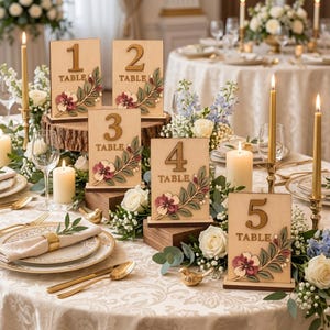 May include: Five rectangular wooden table number signs, numbered 1 through 5, each featuring a laser-cut number and a decorative floral motif with green leaves and maroon flowers. The signs are displayed on wooden blocks on a decorated table.