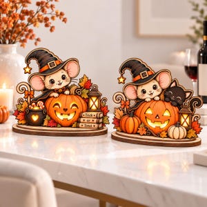 Może przedstawiać: Dwie drewniane dekoracje stołowe na Halloween przedstawiające myszy w czarnych kapeluszach czarownic. Każdy element zawiera świecącą pomarańczową dynię, jesienne liście i małą latarnię. Jeden element zawiera stos książek z napisem Trick or Treat.