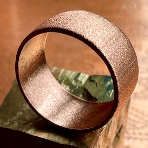 May include: A rose gold-toned ring with a textured, matte exterior surface and a smooth, polished interior band. The ring features a wide, circular band design with a consistent width throughout, showcasing a metallic finish that reflects light.