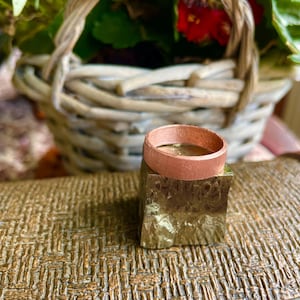 May include: A metallic copper-coloured ring band with a flat, brushed texture resting on top of a small, cubic, gold-toned metallic block that features a rough, crystalline, and faceted surface texture on its sides.