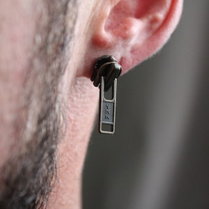 Puede incluir: Un solo pendiente con forma de cremallera, con las letras "YKK" visibles en el tirador. El pendiente es de color metálico oscuro y se lleva en la oreja. El fondo está borroso.