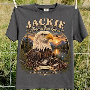 May include: A charcoal gray t-shirt featuring a graphic of a bald eagle in a nest with mountains and a lake. Text reads: JACKIE Forever Our Queen, BIG BEAR LAKE CALIFORNIA, 2012-2026, and FOREVER IN OUR HEARTS. The design includes pine trees and hearts.