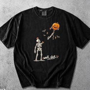 May include: A black short-sleeve t-shirt featuring a graphic of a skeleton missing an arm, with the detached arm floating upward while holding an orange jack-o'-lantern balloon. The text "well, shit..." is printed below the skeleton figure.