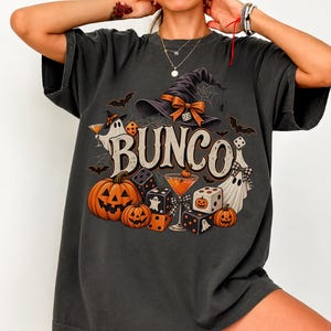 May include: A charcoal gray t-shirt featuring a graphic print with the text BUNCO in large letters. The design includes a witch hat, ghosts, pumpkins, dice, and cocktail glasses in orange, black, and white colors, set against a dark background.