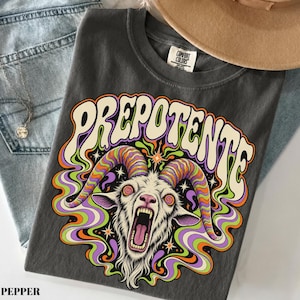 May include: A charcoal gray t-shirt featuring a graphic of a goat with large, multicolored, swirling horns and an open mouth. Above the goat, the word PREPOTENTE is printed in a cream-colored, curved font. The design includes swirling purple, green, and orange smoke-like patterns and small star shapes.