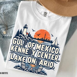 May include: A white t-shirt featuring a graphic print of a lighthouse, a sailboat, and two chairs by the water. The text reads GULF OF MEXICO, KENNEDY CENTER, LAKE ONTARIO. The design includes a sunset, birds, and a city skyline in orange and blue.