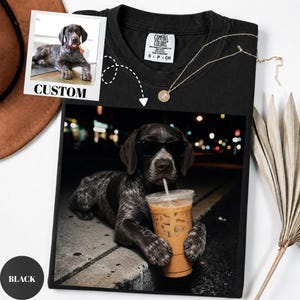 May include: A black t-shirt featuring a graphic print of a German Shorthaired Pointer dog wearing black sunglasses and holding a plastic cup of iced coffee. The shirt includes a small square insert showing a photo of the dog with the text CUSTOM.