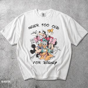 May include: A white short-sleeve t-shirt featuring a colorful graphic of Disney characters including Mickey Mouse, Minnie Mouse, Goofy, Donald Duck, Daisy Duck, and Pluto. The text "NEVER TOO OLD FOR DISNEY" is printed in black above and below the characters.