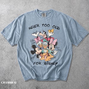 May include: A light blue short-sleeve t-shirt featuring a graphic print of Disney characters including Mickey Mouse, Minnie Mouse, Goofy, Daisy Duck, and Pluto. The text reads NEVER TOO OLD FOR DISNEY, with individual character names written in script.