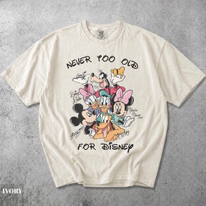May include: An ivory short-sleeve t-shirt featuring a graphic print of Disney characters including Mickey Mouse, Minnie Mouse, Goofy, Donald Duck, Daisy Duck, and Pluto. The text reads NEVER TOO OLD FOR DISNEY with character signatures.