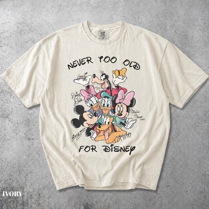 May include: An ivory short-sleeve t-shirt featuring a graphic print of Disney characters including Mickey Mouse, Minnie Mouse, Goofy, Donald Duck, Daisy Duck, and Pluto. The text reads NEVER TOO OLD FOR DISNEY with character signatures.