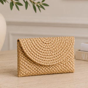 Könnte beinhalten: Eine beige gewebte Clutch mit strukturiertem Flechtmuster und einem geschwungenen Umschlag im Briefumschlag-Stil. Das rechteckige Accessoire besteht aus einem hellbraunen Material mit einem durchgehenden, sich wiederholenden Knotendesign.