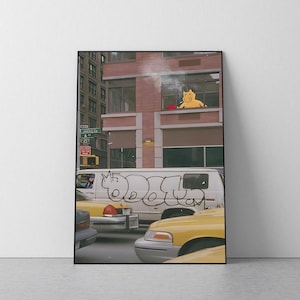 May include: A framed art print depicting a city street scene with a white van featuring black graffiti, yellow taxis, and a red street sign labelled Avenue of the Americas. A yellow cartoon character is visible in a window of a brick building.