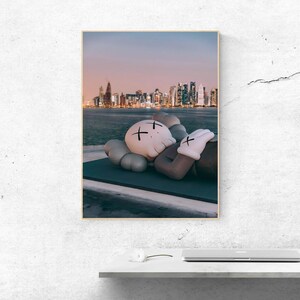 May include: A rectangular wall art print featuring a light-colored cartoon character with X-shaped eyes resting on a dark platform. In the background, a city skyline is visible across a body of water under a soft, gradient sunset sky.
