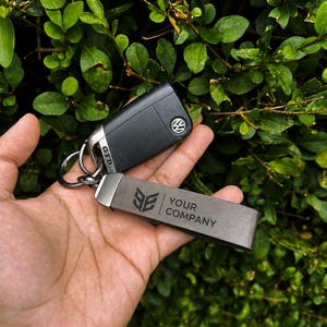 Op de afbeelding: Een zwarte Volkswagen GTD autosleutel bevestigd aan een grijze rechthoekige leren sleutelhanger. De sleutelhanger is voorzien van een donker logo en de tekst YOUR COMPANY in de oppervlakte gegraveerd. Alles zit aan een metalen ring.