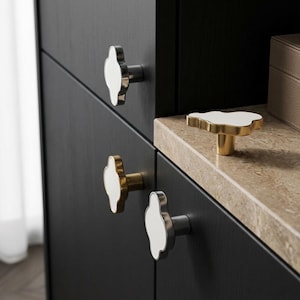 May include: A collection of cloud-shaped cabinet knobs featuring a white top surface and metallic bases in silver and gold finishes. These decorative hardware pieces are mounted on dark wood cabinet doors and placed on a stone countertop.