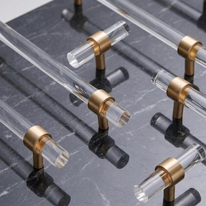 Könnte beinhalten: Eine Sammlung von Schrankgriffen mit klaren, zylindrischen Acrylstangen, die an gebürsteten Messinghalterungen befestigt sind. Die Beschläge sind in einem Raster angeordnet und betonen den Kontrast zwischen dem transparenten Material und dem Metall.