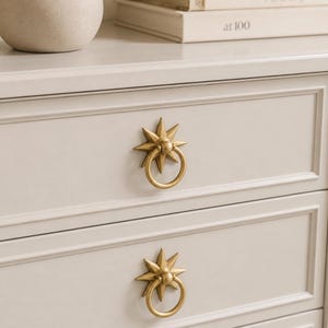 May include: A light gray wooden dresser featuring two drawers, each equipped with a brass-toned star-shaped backplate and a circular ring pull handle. The furniture piece has a smooth, matte finish and clean, rectangular molding on the drawer fronts.