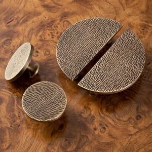 May include: A set of three bronze-toned cabinet hardware pieces featuring a textured, wood-grain-like surface. The collection includes one circular knob shown from the side, one circular knob viewed from above, and one large semi-circular pull handle.