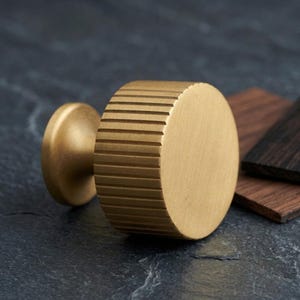 May include: A cylindrical cabinet knob made of brushed brass with a prominent ribbed texture along its side. The knob features a flat circular face and a smaller circular base that connects to the main body, showcasing a metallic gold finish.