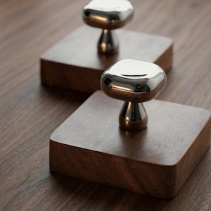 Puede incluir: Dos pomos para armarios con una parte superior de metal plateado pulido y reflectante de forma cuadrada redondeada, unidos a una base metálica cónica. Cada pomo está montado sobre un bloque cuadrado grueso de madera marrón oscura con veta.