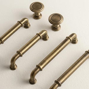 Könnte beinhalten: Ein Set aus antiken Messingbeschlägen, bestehend aus zwei runden Schrankknöpfen und vier zylindrischen Schubladengriffen. Jedes Teil weist eine glatte, gebürstete Metalloberfläche mit einem warmen, gedämpften Goldton auf.