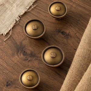 May include: Four circular cabinet knobs with a brushed antique brass finish. Each knob features a domed top, a ridged outer edge, and a small central circular detail embossed with a four-petal flower design. The knobs are arranged on a wooden surface.