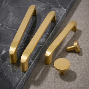 Könnte beinhalten: Eine Sammlung goldfarbener Schrankbeschläge, bestehend aus drei länglichen, bogenförmigen Schubladengriffen und zwei runden Knöpfen. Die Beschläge haben eine glatte, gebürstete Metalloberfläche und liegen auf einer dunkelgrauen Marmoroberfläche.