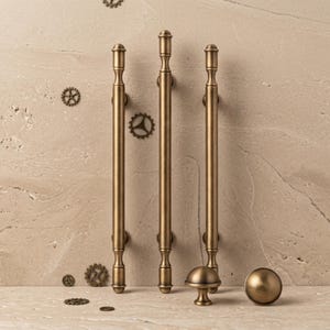 May include: Three vertical antique brass cabinet pulls with a cylindrical rod design and rounded ends, accompanied by two matching round cabinet knobs. The hardware features a brushed metallic finish and is arranged against a textured stone surface.