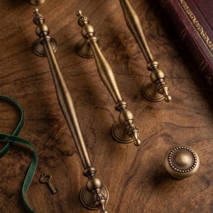 May include: Three antique brass cabinet pulls with a slender, tapered design and decorative spherical accents, alongside a single matching round cabinet knob. The hardware features a brushed metallic finish with detailed, textured circular bases.