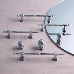 May include: A collection of polished chrome cabinet hardware including three elongated bar pulls with tapered centers and square mounting bases, alongside two spherical knobs featuring horizontal ribbed detailing and circular bases.