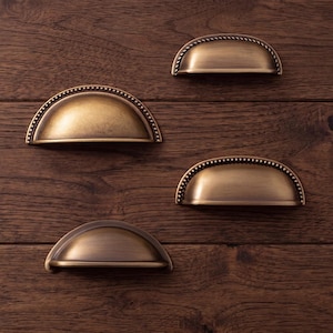 Peut inclure: Quatre poignées de tiroir en métal semi-circulaires avec une finition en laiton antique brossé. Chaque poignée présente un centre bombé lisse et un bord perlé décoratif le long du périmètre supérieur incurvé, offrant un aspect uniforme.