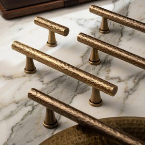 以下が含まれることがあります： ブロンズ調の金属製キャビネットプルハンドル。表面はハンマーで叩いたような質感で、長い円筒形のバーが2つの円錐形の台座で支えられています。全体的に統一された金属仕上げと、繰り返されるくぼみのパターンが特徴です。