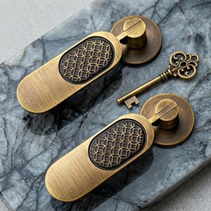 May include: Two antique brass-toned metal drawer pulls with a brushed finish and a central oval inlay featuring a repeating geometric circle pattern. A single ornate brass-toned skeleton key with intricate scrollwork detailing lies between them.