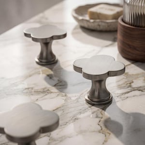 Könnte beinhalten: Drei kleeblattförmige Schrankknöpfe mit gebürstetem Nickel-Finish. Jeder Knopf verfügt über eine glatte, flache Oberseite mit vier abgerundeten Lappen und einen konisch zulaufenden zylindrischen Sockel, der am Befestigungspunkt ansetzt.