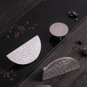 May include: Three silver-toned metal cabinet pulls with a textured, rippled surface. One pull is circular, while the other two are semi-circular in shape. The text "DESIGNED IN MILAN" is visible in small print at the bottom of the image.