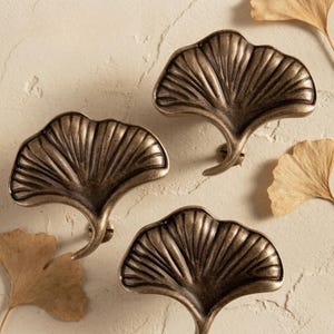 Puede incluir: Tres pomos de metal para armarios con forma de hojas de ginkgo y acabado en bronce oscuro. Cada pomo presenta un diseño de hoja en forma de abanico con nervaduras en relieve que irradian desde el tallo central, creando una textura.