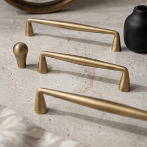 May include: Three brushed brass cabinet pulls with a tapered, elongated design and a single matching round knob. The hardware features a smooth, metallic finish with conical bases that connect to a central bar, providing a clean and functional look.
